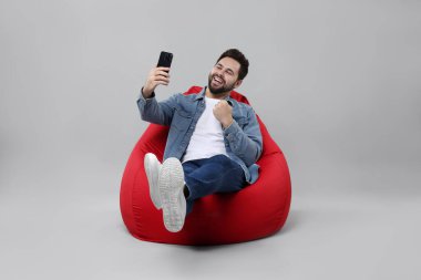 Mutlu genç adam gri arka planda fasulye koltuğu üzerinde akıllı telefon kullanıyor.