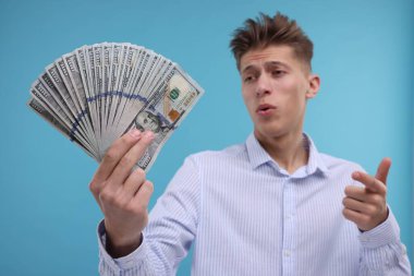 Açık mavi arka planda dolar banknotlarına işaret eden yakışıklı adam, seçici odak