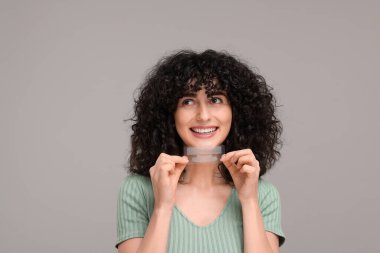 Gri arka planda diş beyazlatma bantları tutan genç bir kadın.