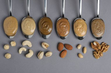 Tasty nut butters in spoons and raw nuts on gray table, flat lay clipart