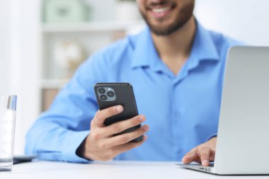Ofisteki beyaz masada bilgisayarla çalışırken akıllı telefon kullanan adam, yakın plan.