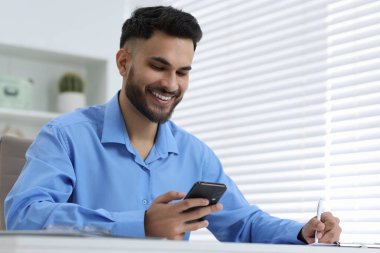 Ofiste beyaz masada çalışırken akıllı telefon kullanan mutlu genç adam, düşük açı