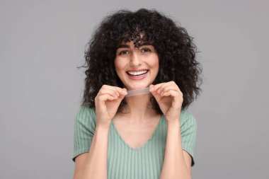 Gri arka planda diş beyazlatma bandı tutan genç bir kadın.