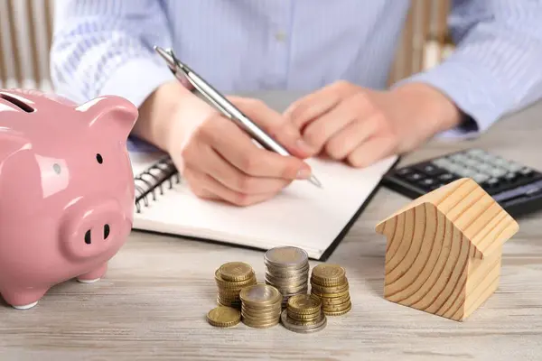 Woman planning budget at wooden table, focus on house model, coins and piggy bank