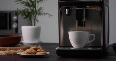 Modern espresso makinesi beyaz fincana kahve dolduruyor. Mutfaktaki siyah ahşap tezgahın üzerine süt dökülüyor.