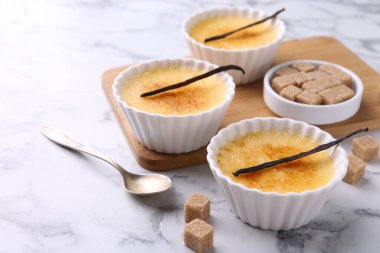 Lezzetli creme brulee kaseler, vanilya kabukları, şeker küpleri ve beyaz mermer masa üzerinde kaşık