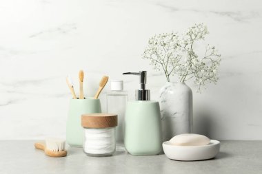 Farklı banyo aksesuarları, kişisel bakım ürünleri ve vazodaki çingene çiçekleri beyaz mermer duvarın yanındaki gri masada.