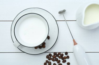 Flat lay composition with mini mixer (milk frother), whipped milk and coffee beans on white wooden table clipart