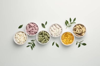 Kâselerde farklı vitamin kapsülleri ve beyaz arka planda yapraklar, düz yatış