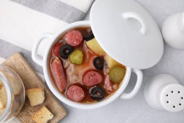 Meat solyanka soup with thin dry smoked sausages in pot on light table, flat lay clipart
