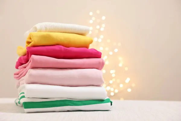 Stack of folded clothes on wooden table against beige background with blurred lights, space for text