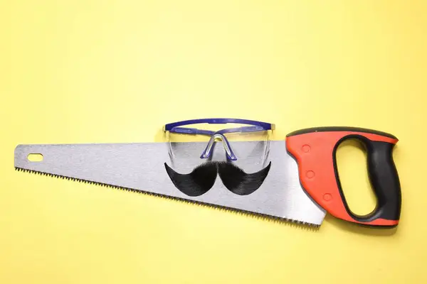 Männergesicht Aus Künstlichem Schnurrbart Schutzbrille Und Handsäge Auf Gelbem Hintergrund — Stockfoto