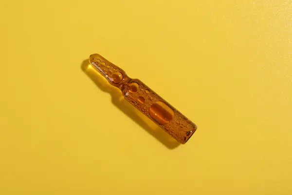 Glass ampoule with liquid on yellow background, top view