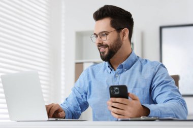 Ofisteki beyaz masada dizüstü bilgisayarla çalışırken akıllı telefon kullanan yakışıklı bir genç.