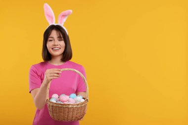 Paskalya kutlaması. Tavşan kulaklı mutlu kadın ve turuncu arka planda boyanmış yumurta dolu hasır sepet, mesaj için yer