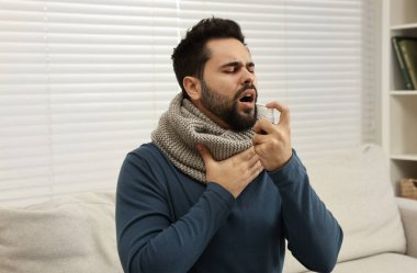 Kapalı alanda boğaz spreyi kullanan atkılı genç bir adam.