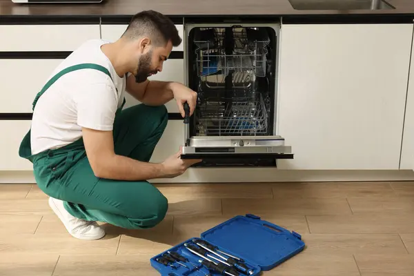 Serviceman repairing dishwasher door with screwdriver in kitchen