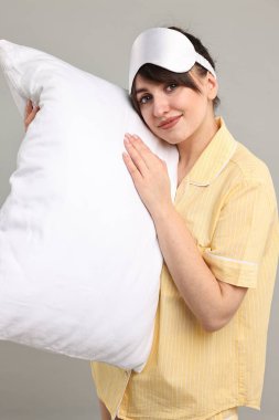 Pijamalı ve uyku maskeli bir kadın gri arka planda yastık tutuyor.