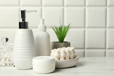 Duvarın yanındaki beyaz masada farklı banyo aksesuarları ve kişisel bakım ürünleri var.