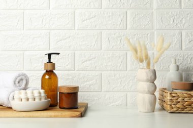 Farklı banyo aksesuarları, kişisel bakım ürünleri ve vazolar tuğla duvarın yanındaki beyaz masada.