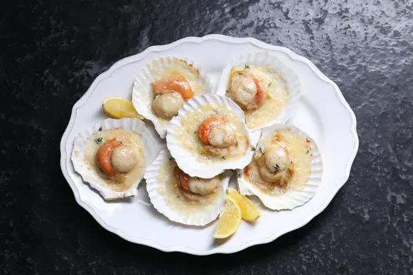 Fried scallops in shells and lemon on black textured table, top view