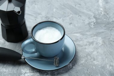 Mini karıştırıcı (süt köpürtücü), fincanda çırpılmış süt ve gri desenli masada moka kabı. Metin için boşluk