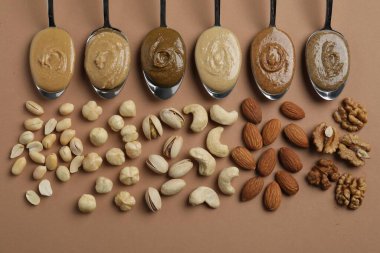 Tasty nut butters in spoons and raw nuts on light brown table, flat lay clipart