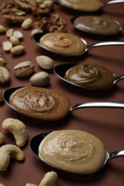 Tasty nut butters in spoons and raw nuts on brown table, closeup clipart