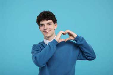 Mutlu genç adam kalp hareketi gösteriyor, elleri açık mavi arka planda.