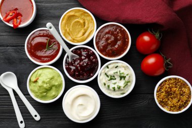 Different tasty sauces in bowls, tomatoes and spoons on black wooden table, flat lay clipart