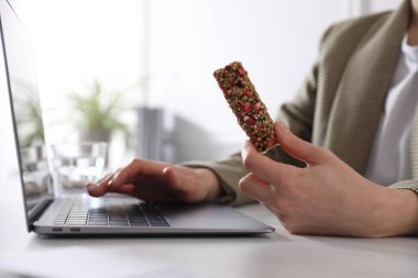Lezzetli granola bar tutan bir kadın ofisteki ışık masasında dizüstü bilgisayarla çalışıyor, yakın plan.