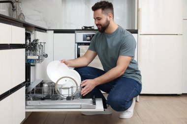 Gülümseyen adam bulaşık makinesini mutfakta tabakla dolduruyor.