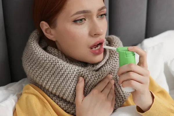 Young woman with scarf using throat spray in bed