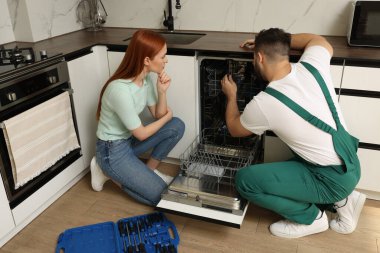 Kadın, tamircinin mutfakta bulaşık makinesini nasıl tamir ettiğine bakıyor.