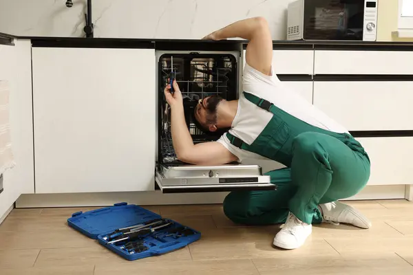 Serviceman repairing dishwasher with screwdriver in kitchen