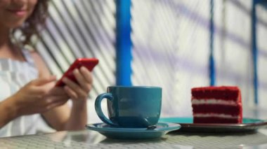 Açık kafede cep telefonu kullanan genç bir kadın, yakın plan. Masada lezzetli kek ve kahve, seçici odaklanma. Kamera sağa hareket ediyor, yavaş çekim etkisi
