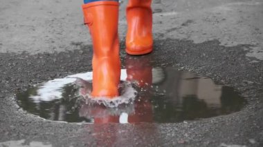 Parlak turuncu çizmeli bir kadın açık havada su birikintisinde yürüyor, yakından bakıyor.
