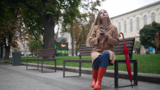 年轻美丽的女人坐在长椅上 在室外用手机 然后下雨时打开雨伞 图库视频片段