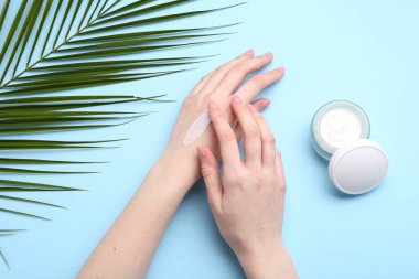 Woman applying hand cream on light blue background, top view