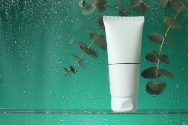 Yeşil zemin üzerinde nemlendirici krem ve okaliptüs dalları olan bir tüp, ıslak cam vasıtasıyla görüş. Metin için boşluk