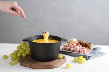 Beyaz ahşap masadaki erimiş peynirle fondü tenceresine bir parça ekmek daldıran kadın. Gri arka planda, yakın plan.