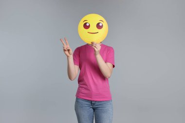 Gülümseyen ifade ile yüzünü kapatan ve gri arka planda barış işareti gösteren kadın