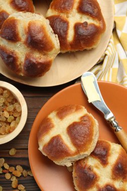 Lezzetli haçlı çörekler, tereyağlı bıçak ve kuru üzümler ahşap masada, düz uzanmış.
