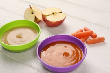 Baby food. Purees of apples and carrots in bowls on white wooden table clipart