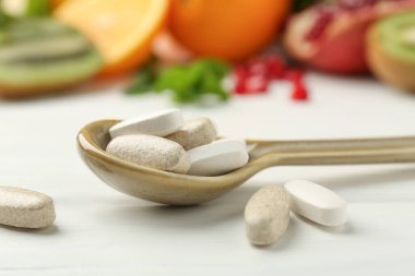 Farklı vitamin hapları kaşıkta ve taze meyveler beyaz ahşap masada, yakın plan.