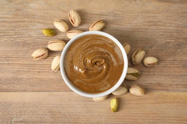 Delicious nut butter in bowl and pistachios on wooden table, top view clipart