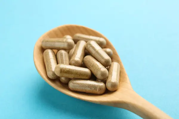 Vitamin capsules in wooden spoon on light blue background, closeup