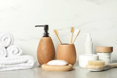 Beyaz mermer duvarın yanındaki gri masada farklı banyo aksesuarları ve kişisel bakım ürünleri var.