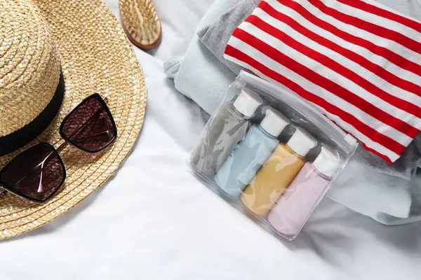 Cosmetic travel kit. Plastic bag with small containers of personal care products, beach accessories and stack of clothes on bed