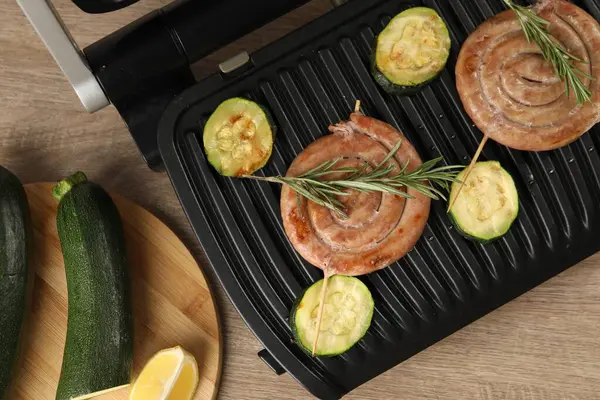 Electric grill with homemade sausages, zucchini and rosemary on wooden table, flat lay